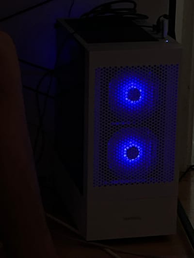 The image shows a white computer tower with two blue illuminated fans on its top. The computer is sitting on a dark surface, likely a desk, and is angled towards the viewer. Cables are visible behind and beneath the computer, suggesting it is connected and in use. The lighting is dim, with the blue glow from the fans being the primary light source in the immediate vicinity of the computer. In the foreground, a person's arm and leg are visible, indicating someone is sitting or positioned near the computer. The background is dark and indistinct, providing little information about the room's details. A small white object with a blue indicator light is attached to the left side of the computer's top. The overall impression is of a gaming or computer setup in a dimly lit room.