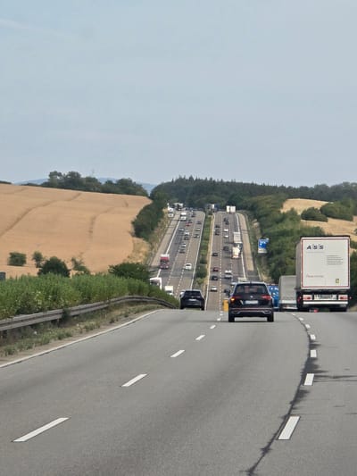 The image captures a multi-lane highway in Dauborn, Germany, under a clear sky. The highway curves and ascends into the distance, lined with trees and a golden-hued field on the left. Traffic is moderate, with numerous trucks and cars traveling in both directions. One prominent truck, with the "ASS Transports" logo and "Schmitz" branding, is visible in the right lane. Several other trucks and vehicles are scattered throughout the lanes. The road surface is asphalt with clear white lane markings. A metal guardrail runs along the left side of the highway, separating it from the embankment. The overall scene depicts a typical highway scene with ongoing traffic flow in a rural setting.