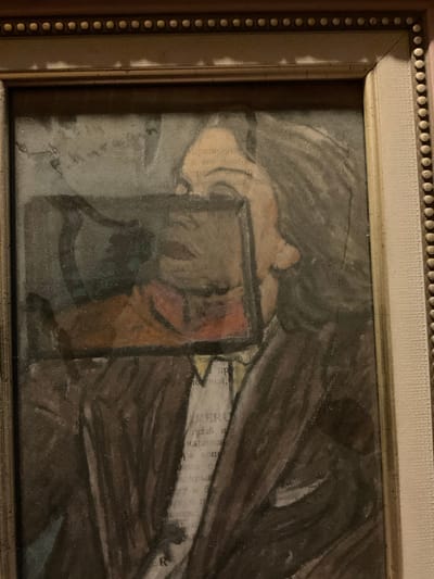 This is a close-up, dusty, framed image of a painting. The painting depicts a person, possibly a child, lying down. A framed object, possibly a picture or a small mirror, is held up near the person's face. The background is dark and indistinct, with swirling brushstrokes suggesting drapery or shadows. There are fragments of text visible on the painting, appearing to be from a newspaper or book, with some Cyrillic letters discernible, such as "АР" and "д", "и". The style of the painting is impressionistic or expressionistic, with visible texture and layering of colors, including muted browns, grays, and a touch of orange in the framed object. The lighting in the photograph is dim, casting shadows and highlighting the dust on the surface of the artwork. The frame is light-colored with a beaded border. Given the location context of Sergeevo, Russia, the text is likely in Russian. The overall mood is somber and reflective.