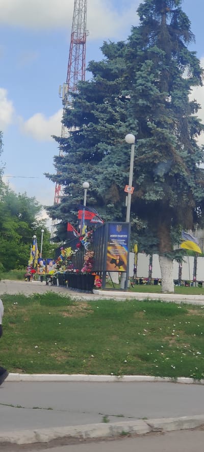 The image depicts a memorial site in Valky, Ukraine. A tall telecommunications tower is visible in the background, partially obscured by a large evergreen tree. In the foreground, a black metal fence serves as the backdrop for a memorial display. The fence is adorned with numerous colorful flowers, flags, and framed photographs of individuals. A prominent sign displays the Ukrainian trident emblem and text in Ukrainian, which translates to "Avenue of Memory". Another sign features a candle and an image of Ukraine. Ukrainian flags and various other banners are also present, some with text that is partially legible, such as "KRAKEN". The memorial appears to be situated on a grassy area with a paved walkway in front of it. The sky is partly cloudy. The overall scene is solemn and suggests a tribute to fallen individuals.