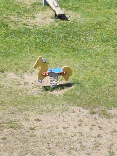 The image shows a yellow, spring-mounted toy horse on a playground. The horse is a classic design with a blue seat and red accents. It is positioned on a patch of sand surrounded by grass. In the background, a portion of a wooden seesaw is visible. The scene appears to be outdoors, under natural lighting, suggesting a daytime setting. The location is identified as Gardene, Latvia.