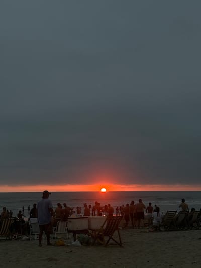 The image shows a large group of people on the beach in Montañita, Ecuador (latitude: -1.825, longitude: -80.754). The sun is setting on the horizon, casting an orange glow over the sky and the ocean. There are many people in the distance, standing in the water and on the sand, and some people sitting in beach chairs in the foreground. There are also several coolers and some trash on the sand. The beach is sandy and there are some small waves in the ocean. The environment is a tropical beach setting. The people appear to be enjoying the sunset and the end of their day.
