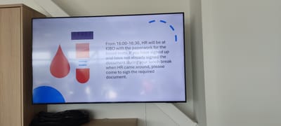 A medium shot captures a television mounted on a wall displaying a message. The TV screen shows a graphic with stylized blood drop imagery and the text: "From 16:00-16:30, HR will be at KIBO with the paperwork for the blood tests. If you have signed up and have not already signed the document during your lunch break when HR came around, please come to sign the required document." The location is Λάκκωμα, Greece. Below the TV is a shelf or platform, upon which rests a dark-colored, crumpled object. The surrounding environment includes a light-colored wall and a section of a wooden cabinet adjacent to the TV. The scene appears to be indoors, possibly within a workplace or office setting.