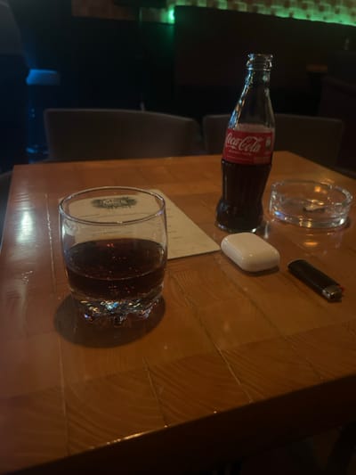 A table with a partially filled glass of cola sits in the foreground. Next to it, a menu card is visible. Further up, a Coca-Cola bottle, a white earbud case, an ashtray, and a lighter complete the collection of items. The table appears to be made of wood, and the image is taken in a dimly lit setting. Several chairs are also visible in the blurred background.