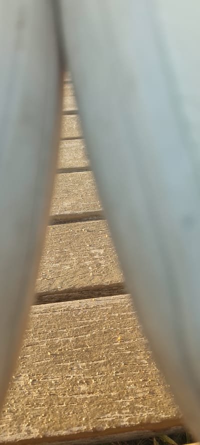 The image shows a close-up view of wooden planks, possibly part of a walkway or bench, seen through a narrow gap between white or light-colored surfaces. The wood is textured with visible grains and appears to be weathered or aged. Some small light-colored particles are visible on the wooden surface. There are grass blades or weeds in the upper right. The image does not provide enough information to determine the location in Nahariyya, Israel. There is no indication of people, actions, or interactions in the picture.