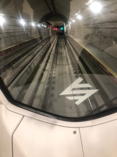 A view from inside a train carriage looking down a subway tunnel. The tunnel walls are concrete and lit by rows of lights. In the distance, a green light is visible. The foreground shows a train window with a white VIA Rail logo on it. Reflections of the tunnel and tracks appear on the glass. A hint of the white interior of the train is visible around the window.
