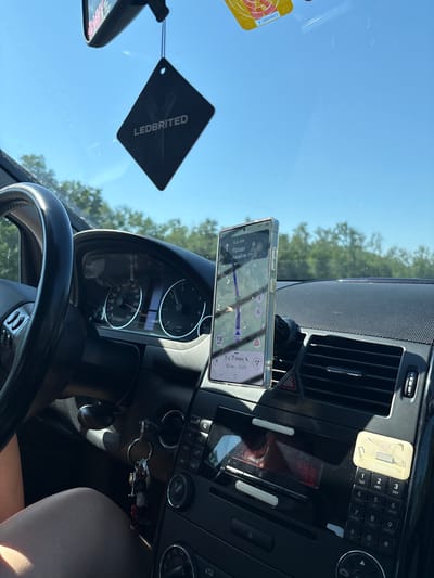 The image shows the interior of a car with a navigation system displayed on a smartphone mounted on the dashboard. The phone is displaying a route with a distance of 6.6 km and an estimated arrival time of 1 hour and 7 minutes. The car's dashboard includes a steering wheel, instrument cluster, and audio system controls. The view outside the car through the windshield shows trees and a clear blue sky. There is a car air freshener hanging from the rearview mirror. A person's leg is visible in the lower left corner of the image. The context suggests a road trip or commute. Given the visible navigation prompts in Cyrillic script ("Продо лжайте де"), and the geographical context of Ruma Municipality, Serbia, this scene likely depicts a journey within or near this region.