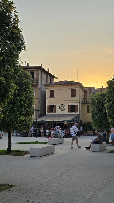This is a vertically oriented image of a street scene in Peschiera del Garda, Italy, at sunset. The sky is a gradient of pale yellow and orange, indicating the sun is low on the horizon. In the foreground, a tree with lush green foliage stands to the left. A paved walkway with a textured surface extends into the distance, lined with modern, light-colored concrete benches.

Several people are visible in the scene. Some are sitting on the benches, while others are walking or standing. One man in a white shirt with a pattern is walking across the walkway. Further back, a building with a light-colored facade features brown shutters on its windows and a sign with the text "GTS TRO" in a circular logo. There are tables and chairs set up outside what appears to be a restaurant or cafe, with more people seated there. The overall atmosphere suggests a relaxed evening gathering. In the background, more buildings and trees are visible, and a few television antennas are perched on rooftops.