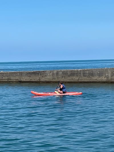 In the city of Khostinskiy, Russia, a person is paddleboarding on the Black Sea. The person is wearing a life vest and is seated on a red paddleboard. The paddleboard is on the water, and the person is holding a paddle. In the background, there is a concrete barrier and the ocean. The sky is blue and clear.
