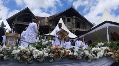 A group of men stand in front of a white tent in a compound in Rukoko, Democratic Republic of Congo. They are wearing white robes and hats. In the center, a man holds a leopard skin. Several men are seated on chairs behind them. In the foreground, a row of white and peach flowers line the front of the tent. A man is standing behind a wooden podium decorated with two golden crests. Trees are visible behind the group. The building behind the group appears to be under construction. The sky is partly cloudy.