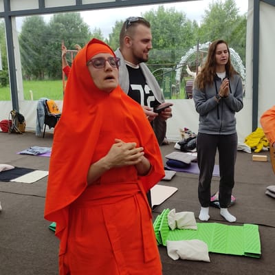 The image depicts an outdoor gathering under a tent, possibly for a yoga or meditation session, at the village of Zhirnosovo, Russia. In the foreground, a person wearing an orange robe and glasses with their eyes closed is standing with hands clasped at their chest. Behind them, a man in a black t-shirt and grey cardigan looks to the side, holding a smartphone. To the right, a woman in a grey hooded jacket stands with her hands clasped in front of her chest, her eyes also closed. Several yoga mats and cushions are scattered on the ground. In the background, trees and a cloudy sky are visible, suggesting it is daytime. The overall atmosphere appears calm and serene.