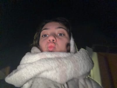 An upside-down selfie shows a person with fair skin, dark eyes, and short brown hair. They are wearing a fluffy, light-colored robe or hoodie around their head. The person is making a pouting expression with their lips. The background is dark and indistinct. A hint of a wooden surface is visible in the upper left corner. The image is somewhat grainy.