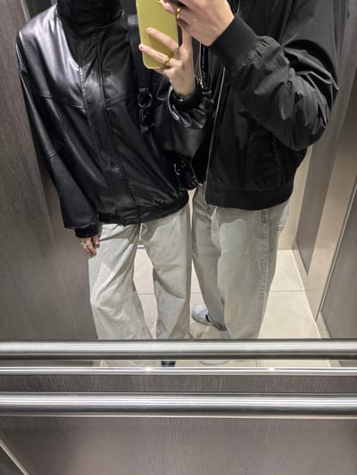 An indoor photograph, presumably a mirror selfie, depicts two individuals standing reflected in a mirror, likely inside an elevator in Cachan, France. Both subjects are partially visible from the chest down, dressed in casual attire. The individual positioned on the left wears a black leather-like jacket with a high collar and light-colored, relaxed-fit jeans. A ring is visible on their left hand, which is holding a light-yellow smartphone, positioned to capture the image. A black strap, likely from a shoulder bag, is visible across their left side. The individual on the right wears a black bomber jacket with ribbed cuffs and hem, light-colored, relaxed-fit jeans, and white sneakers with black side stripes. A ring is visible on their right hand. The elevator interior features light brown wood-grain paneled walls and a light-colored tiled floor. Two parallel stainless steel handrails span the lower foreground of the reflective surface.