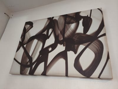 The image shows a close-up, angled view of a canvas artwork with abstract dark brown brushstrokes on a white background. The artwork is mounted on a white wall. In the upper left corner of the image, part of the ceiling is visible, showing some small dark marks and a small hole with a wire. The overall scene appears to be an indoor setting. There is no visible indication of the specific location within Cavite City, Philippines.