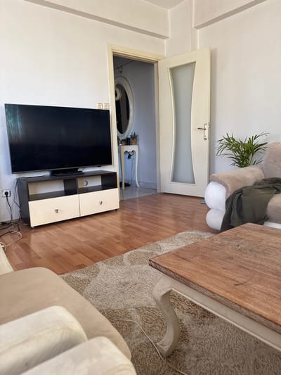 The image depicts a living room scene, likely within an apartment in Büyükçekmece, Turkey. In the foreground, a wooden coffee table with an ornate white base sits on a shaggy, beige rug. To the right, a portion of a cream-colored sofa is visible, with a dark green garment draped over its armrest. Behind the sofa, a potted plant with green fronds adds a touch of nature to the room.
Dominating the left side of the frame is a large, black flat-screen television set perched atop a two-tiered media console. The console has a dark brown top and frame, with two cream-colored drawers at the bottom, each adorned with a silver knob. Electrical cords are visible connected to the television and the wall socket.
In the background, an open doorway leads into another room. Through the doorway, a hallway is partially visible, with a decorative white table and an oval mirror on the wall. Further down the hallway, another door with frosted glass and a silver handle is slightly ajar. The walls of the living room are painted a light, neutral color, and the floor is covered in polished wooden planks. The overall impression is one of a comfortable, lived-in space.