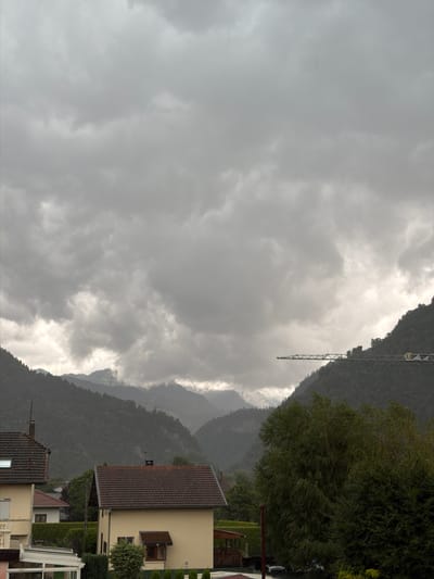 This is a vertically oriented, wide-angle photograph of a cloudy, overcast day in a mountainous region, likely Cluses, France. The scene is divided into distinct areas: the sky, the mountains, and some foreground buildings.
The sky dominates the left side of the image, filled with heavy, grey clouds that obscure much of the light. Towards the right and background, the clouds break slightly, revealing glimpses of the mountainous landscape behind. The mountains themselves are covered in dense, dark green vegetation, appearing somewhat muted by the weather. A tall, skeletal construction crane is visible against the sky and mountains on the far right, suggesting ongoing construction or a former industrial site.
In the foreground, on the right side of the frame, are parts of residential buildings. One building is prominent, with a cream-colored facade and a dark-tiled roof. It features a small balcony with a railing and a window. Another building, partially visible, also has a dark-tiled roof. The architecture suggests a European setting. Trees and hedges are also present, contributing to the residential feel.
The weather appears to be stormy or very overcast, with diffused light and a generally somber atmosphere. The time of day is not precisely discernible due to the cloud cover, but it is likely daytime. There is no visible text in the image. The overall impression is one of a serene, albeit cloudy, mountain village setting.