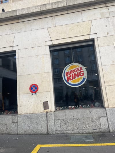 The image captures the exterior of a Burger King restaurant on the ground floor of a building in an urban environment, identified as Genève, Switzerland. The primary subject is a large rectangular window featuring the iconic circular Burger King logo. The logo displays "BURGER KING" in red and yellow text, set against a blue and yellow bun-shaped graphic.
The building's facade is constructed from light-colored, possibly beige or off-white, cut stone or large tiles, giving it a structured appearance. The window reflects the architecture of an opposite building, showing its own windows and facade details.
Below the main Burger King window, a small red and blue circular traffic sign is affixed to the stone wall, indicating "no stopping" or "no standing" (a red cross on a blue background). Directly beneath it is a small white rectangular sign with a black double-headed arrow, which typically denotes the zone of the restriction.
To the right of the building, a grey, granular sidewalk or pavement made of stone blocks leads to an asphalt street. A clear yellow line is painted along the edge of the street, and a metal grate is embedded in the pavement. The lighting suggests it is daytime, with diffused light indicating an overcast sky or an indirect light source, as there are no distinct shadows. The scene portrays a commercial storefront on a typical city street.