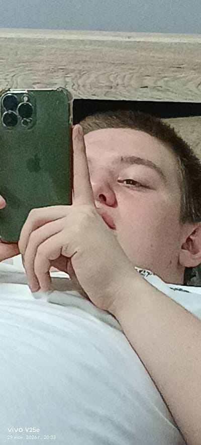 A selfie is taken by a person holding a green iPhone in their left hand. The person's right index finger is held to their lips, suggesting they are asking for silence. The background appears to be a wooden headboard. The timestamp "29 июня. 2026 г., 20:33" and "vivo V25e" are visible in the bottom left corner, indicating the photo was taken on June 29, 2026, at 8:33 PM. There is no visible indication of the city of Семилуки, Russia.