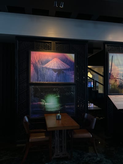 Indoor scene with a table and two chairs set against a wall decorated with artwork. The artwork consists of framed pieces with ornate detailing. The table is set with a small potted plant. The location is Nusa Dua, Indonesia. No people are visible.