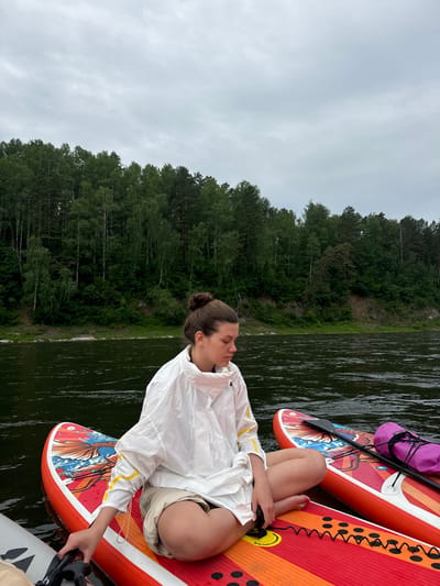 The media file shows a woman sitting cross-legged on an orange and red paddleboard. She is wearing a white hooded jacket and beige shorts. Her eyes are closed, and she appears relaxed, possibly meditating or enjoying a moment of peace. The paddleboard has a colorful design featuring a fish and waves.
The paddleboard is on dark, rippling water, and another paddleboard is visible behind it, with a purple bag attached. The background features a dense, green forest that rises from a rocky bank along the water's edge. The sky is overcast and cloudy, suggesting a temperate climate. The overall scene evokes a sense of tranquility and connection with nature.
The image was taken in Манский, Russia. There is no visible text in the image. The time of day appears to be daytime, but the cloudy weather might obscure direct sunlight.