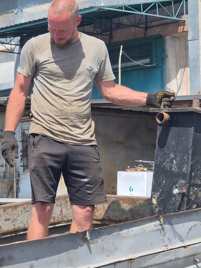 A bald, fair-skinned man with a beard and a light-colored t-shirt and dark shorts stands in a weathered metal container. He wears black work gloves with yellow accents on his right hand, which is resting on the edge of another metal structure. His legs are visible up to his knees, and he appears to be inside the container. Behind him, a white refrigerator with a blue logo is visible, along with other assorted items and structures that suggest an industrial or outdoor work environment. The scene is lit by sunlight, casting shadows. The man is in Pidhaitsi, Ukraine.