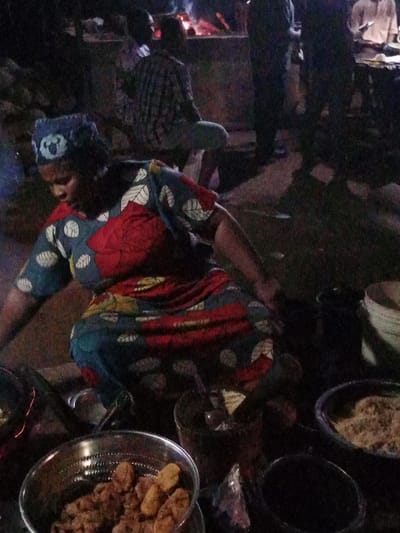 This image depicts a nighttime scene in Ube, Nigeria, centering on a woman engaged in food preparation or vending. A dark-skinned woman, wearing a patterned dress featuring blue, red, and yellow with white leaf motifs, and a blue patterned headwrap, is crouched low on the ground. In front of her, to the lower left, is a metallic bowl containing golden-brown fried food items, possibly plantain or fritters. To her right, a mortar and pestle are visible next to a dark bucket, and further right, a dark bowl holds a light-colored granular substance. Behind these, a white bucket is partially visible. To the woman's immediate left, a cooking pan is present, emitting a faint glow. In the mid-ground, a blurred individual in a plaid shirt is seated on a low surface, facing a visible fire with glowing embers. Other indistinct, blurred figures are present in the dark background. The ground appears to be unpaved dirt or a rough surface. The overall scene is dimly lit by artificial light sources, including the cooking fire.