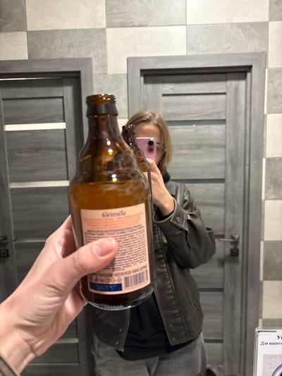 A person in a brown leather jacket is holding a brown glass bottle labeled "Grindle" in front of a mirror. The person is reflected in the mirror, taking a photo with a pink phone. The background includes a tiled wall and a gray wooden door with vertical grooves. The scene is presumably inside a bathroom based on the tiled wall and mirror. There is no explicit information to conclude the location as Ust-Ilimsk, Russia.