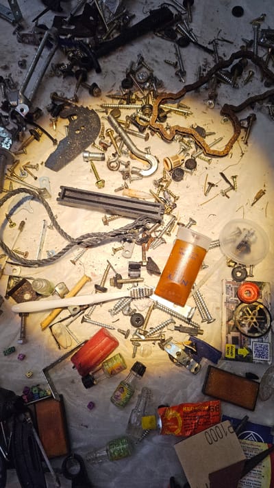 The image presents a top-down view of a cluttered, light-colored surface, possibly a workbench or floor, illuminated by a direct, warm light source that creates distinct shadows. The scene is filled with a miscellaneous collection of small objects, indicating a setting like a workshop or storage area, likely in Cam Đức, Vietnam.
The primary subjects are numerous metal fasteners, including a wide variety of screws (phillips, slotted, hex head), nuts, bolts, and washers, many showing signs of wear or rust. Other hardware items include metal brackets, a spring, a U-shaped hook, a braided metal cable, and a rusty, wavy chain-like component or gasket.
Among the clutter are several containers and personal items. A clear plastic container with a lid displays "NSX 03/04/2025" and "HSD: 28/04/2026". An orange cylindrical bottle is partially labeled "HAND WASH" and "NYV". A red tube, likely an adhesive, shows "KEO 56", "Keo", and "VANG". Several miniature glass bottles with green labels and black caps are scattered across the surface. Other items include a white toothbrush, multiple small, colorful dice (purple, green, orange), and a piece of sandpaper marked "P1000".
Visible text on a piece of paper includes a "Google Maps" logo, a QR code, and extensive Vietnamese text: "29", "02583.823.999", "Phòng Hàng Số", "TẠI NINH HO", "822 Trần", "Số 8, Hồng", "2. Mương Than", "318 564", "37 Nguyễn Văn Cư", and "Nhận hợp đồng thai". A stylized Yamaha logo is also partially visible on another item. No specific activity or event is taking place; the scene suggests a collection of assorted items, possibly awaiting organization or disposal.