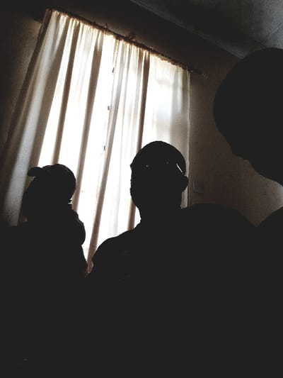 The scene depicts an indoor location in Panhauya, Nigeria. Three human figures are present, rendered in deep silhouette against a brightly lit window centrally positioned in the background. The window is covered by light-colored, pleated curtains, suspended from a dark curtain rod. From left to right, the first figure is partially obscured and appears to be wearing a cap or head covering. The central figure is fully silhouetted, facing the viewer's general direction, with discernible outlines suggesting eyeglasses. The third figure, on the right, is visible from the side, presenting a profile of their head and upper torso, facing towards the right edge of the frame. The remainder of the room, including the ceiling and walls, is underexposed, revealing minimal detail. No specific actions or interactions are discernible among the individuals.
