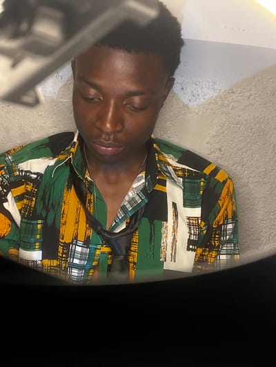 The image depicts a young adult Black male, centrally positioned in an interior setting in Bar Gada, Nigeria. He is shown from the chest up, with his head slightly bowed and eyes directed downwards. He wears a long-sleeved, collared button-down shirt featuring an abstract pattern of vertical and horizontal brushstrokes and grid motifs in shades of green, yellow, black, and white. A thin, silver-colored ball chain necklace is visible around his neck, beneath an untied black tie draped loosely around his collar. The background consists of a light-colored, textured wall, possibly stucco. An out-of-focus, dark gray object, appearing metallic or plastic, occupies the upper left portion of the frame, possibly a light fixture or a photographic accessory. The lower section of the image is obscured by a dark, out-of-focus foreground element. The subject is still, with no apparent action or interaction with others.