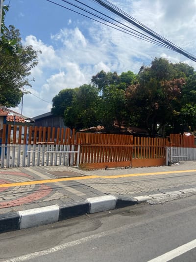 The image shows a street in Mojokerto, Indonesia (latitude: -7.481, longitude: 112.44). The street is paved with asphalt and has a sidewalk with a yellow painted edge. The sidewalk is made of concrete blocks and a curb. There is a wooden fence with orange slats on the right side of the image. The fence is partially open. In the background, there is a group of trees and a blue sky with white clouds. There are multiple electrical wires running across the sky.