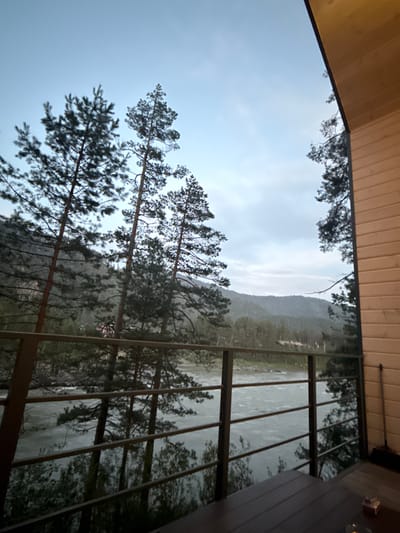 The image depicts a view from a wooden balcony overlooking a river and a forested mountain landscape. Tall pine trees are visible in the foreground and midground. The sky is overcast and the light is subdued, suggesting it may be early morning or late evening. The river is wide and appears to be flowing quickly, with ripples on its surface. In the background, a range of mountains covered in dense forest stretches across the horizon. On the right side of the frame, the wooden exterior of a building is visible, along with a broom leaning against the wall. The location is identified as Barangol, Russia.
