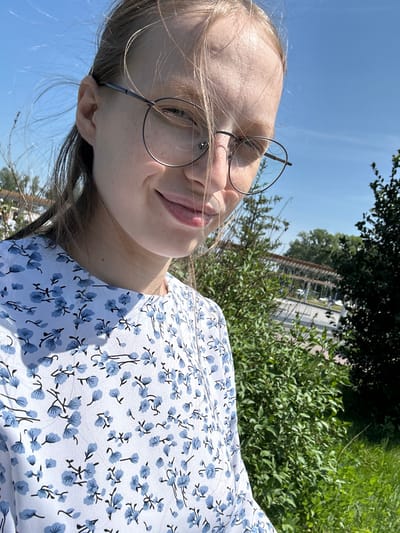 A young woman with blonde hair and glasses is taking a selfie outdoors. She is wearing a white dress with a blue floral pattern. Her hair is blowing in the wind, partially obscuring her face. Behind her, there are green trees and bushes, and a bright blue sky. In the background, there is a structure with a wooden roof and some cars. The scene appears to be in a park or natural area within the city of Металлплощадка, Russia.