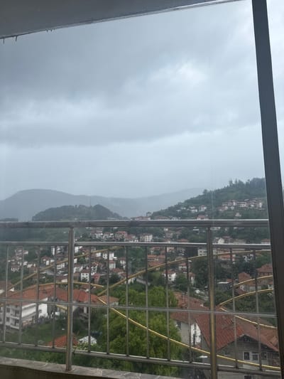 The image displays a view of a village nestled in a hilly landscape, likely Momchilovtsi, Bulgaria, on a cloudy day. The foreground features a metallic railing with vertical bars and horizontal support elements. Beyond the railing, a cluster of houses with red-tiled roofs are visible, interspersed with trees. The houses are built on a sloping terrain, with some appearing to be multi-story. Further in the distance, more houses extend up the hillsides. The background consists of layers of mountains and hills shrouded in mist or clouds, creating a subdued and atmospheric scene. The overall impression is that of a rural or semi-urban settlement in a mountainous region. There are no visible people or specific actions depicted in the image. The weather appears overcast, possibly indicating rain or damp conditions.