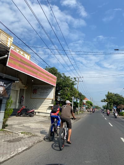 Two men are riding bicycles down a paved road in Kecamatan Tanggulangin, Indonesia, located at latitude: -7.497, longitude: 112.686. The man in front is wearing a brown t-shirt and a white cap. The man behind him is wearing a blue and red shirt and a black backpack.  There are many power lines overhead and several buildings lining the road. The sky is mostly blue with some white clouds. Other vehicles can be seen in the distance, including a motorbike and a few cars. There are several small trees along the street and some greenery in the background. The men are riding in the same direction, and there are no other people visible. The road is relatively wide, with no obstacles present. The ground is paved with a light grey asphalt. The day is clear and sunny.