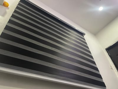 The image depicts a closed, gray and black striped window shade hanging in a room. The location is not discernible from the image and cannot be placed in Iddo Tudunwada, Nigeria. The shade appears to be made of fabric, and the stripes alternate between gray and black. The shade is mounted inside the window frame and the bottom of the shade has a white border. The room has white walls and a light fixture in the ceiling. A portion of a door is visible on the right side of the image.