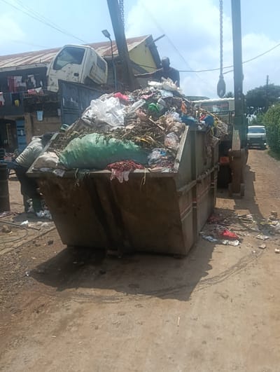The image shows an overflowing garbage container on a dirt road in Muguga Ward, Kenya. The container is filled with a mix of waste materials including plastics, textiles, and organic debris. A sanitation worker stands in the container, adjusting the load. The container is attached to a waste management vehicle, with another man on top. The scene is cluttered, with refuse scattered around the base of the container on the ground. Buildings and power lines are visible in the background.