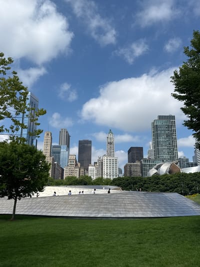 The image depicts a sunny day in Chicago, United States of America. The scene is a cityscape with skyscrapers visible in the background under a clear blue sky dotted with white clouds. In the foreground, there's a large, curved, polished metal structure with a reflective surface. Adjacent to this structure is a vibrant green lawn. Several people are seen on and around the metal structure, appearing to be enjoying the outdoors and interacting with the artwork. Trees with lush green leaves frame the scene, adding a touch of nature to the urban environment. The time of day appears to be midday, given the bright sunlight casting shadows. The overall atmosphere is one of leisure and appreciation for public art and green spaces within a bustling city.