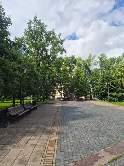 This is a vertical, eye-level, outdoor shot of a park pathway in Petrozavodsk, Russia. The pathway is paved with grey cobblestones and bordered by a reddish-pink stone edge and green grass. The pathway leads the eye toward a lush green park filled with large trees and foliage. A dark-colored park bench with wooden slats sits on the grass beside the pathway. In the background, a statue stands on a stone pedestal, surrounded by trees. Two black lampposts are also visible within the park. The sky above is partially cloudy, with patches of blue sky peeking through. The overall lighting suggests it is daytime, possibly late afternoon or early morning, with soft sunlight filtering through the trees. There is no visible text in the image. The scene evokes a sense of peacefulness and urban nature.