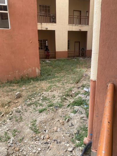 The image depicts an outdoor, undeveloped ground area situated between two multi-story buildings in Baganakwo, Nigeria. In the foreground, the left side shows a portion of a reddish-brown building wall with a white-framed window at the upper left. The wall exhibits fading and minor disfiguration. The right foreground reveals part of another reddish-brown building wall, distinguished by a cream-colored vertical trim element. Two prominent orange-brown pipes are mounted vertically on this wall, with a black cable emerging from the ground near their base.
The central ground area consists of dry, uneven earth with sparse green vegetation, primarily weeds and grass, interspersed with scattered debris including small rocks, concrete fragments, and plastic waste.
In the background, a two-story cream-colored building is visible, featuring recessed balconies or walkways with brown railings on both levels. On the lower left balcony, a person is standing, largely obscured from the waist down by the railing. The individual wears dark upper attire and what appears to be red or patterned lower clothing, facing away from the camera and looking towards the left. Behind them is a window with multiple panes. The upper left balcony also has a multiple-paned window and a railing. The lower right balcony is vacant but features a closed door and an external air conditioning unit mounted on the wall below the railing. The upper right balcony section shows a closed door and railing. The scene is brightly lit by natural light.