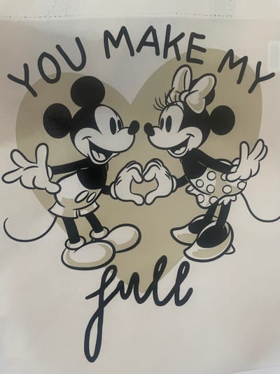 This is a close-up, eye-level, medium shot of a white tote bag with a printed design. The design features cartoon characters Mickey Mouse and Minnie Mouse in black and white, positioned in front of a large, light brown heart. Both characters are smiling and facing each other, with their hands forming a heart shape between them. Above them, the words "YOU MAKE MY" are printed in black, stylized block letters. Below the characters, the word "full" is printed in black, cursive script. The overall image is well-lit, with no shadows obscuring the details of the print. The location is not discernible from the image itself.