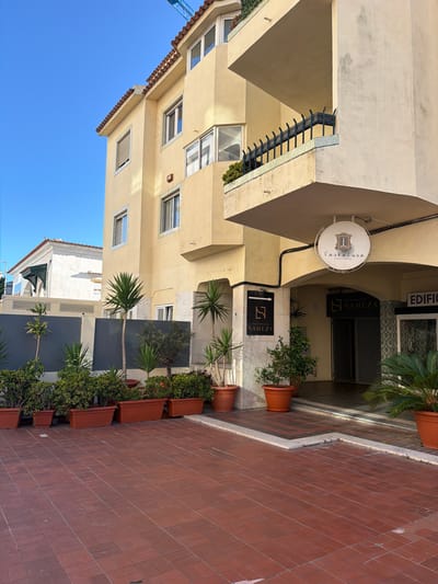 The image depicts the exterior of a building in Cascais, Portugal, under a clear blue sky. The building is a multi-story structure painted a pale yellow. It features a red tile roof and several windows, some with white frames and others with dark trim. A prominent feature is a large balcony or overhang with a black metal railing.
At street level, an arched entryway leads into the building. Above the entrance, a circular sign displays the name "TascaLusa" with an illustration of a window and roof. To the left of the entrance, a black rectangular sign with a white logo and the word "NAMEZA" is mounted on the wall, along with a white arrow pointing to the right. Another sign to the right of the entrance reads "EDIFICIO".
The area in front of the building is paved with reddish-brown rectangular tiles. Several potted plants are arranged along the front of the building, including palm-like plants and dense green shrubs. A darker gray fence or wall is visible behind some of the plants. In the background, a white building with a green awning and arched windows can be seen, as well as a portion of a blue construction crane. No people are visible in the scene.