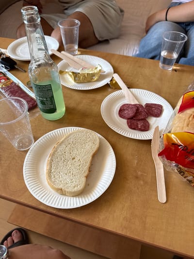 In Sveti Vlas, Bulgaria, a wooden table is laid out with food and drinks, possibly for a casual gathering. On the table, there are several paper plates. One plate holds a slice of white bread. Another plate displays a few slices of salami, accompanied by a wooden knife. A third paper plate, partially wrapped in gold foil, contains what appears to be a small dessert or pastry with frosting.
A clear glass bottle with a green liquid, likely a beverage, stands upright, its label partially visible. Next to it are two clear plastic cups, one filled with a clear liquid, possibly water, and another empty cup. A set of keys is also present on the table.
In the background, two individuals are seated. One person is wearing blue jeans and a black bracelet on their wrist. The other person is wearing light-colored shorts and a light-colored top. The environment appears to be indoors, with hints of furniture and a relaxed atmosphere. The overall scene suggests a shared meal or snack.