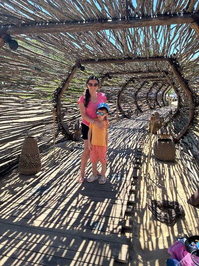 In the city of Shkorpilovtsi, Bulgaria, two individuals, a woman and a young boy, are standing on a wooden boardwalk beneath a tunnel-like structure constructed from interwoven branches and logs. The woman, on the left, wears a pink t-shirt and black shorts, with sunglasses and her hair pulled back in a ponytail. The boy, on the right, is wearing an orange t-shirt, pink shorts, a blue baseball cap, and sunglasses. He is positioned slightly in front of the woman, who has her arm around his shoulders.
The structure creates a dappled effect on the boardwalk and sandy ground due to the sunlight filtering through the branches. Various objects are present within the scene, including woven baskets, a torch-like decoration on a stick, and a heart-shaped bundle of twigs lying on the sand. In the background, through the opening of the branch structure, a parked vehicle is visible. The overall environment appears to be outdoors, likely a scenic spot in Shkorpilovtsi.