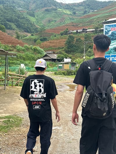 Two males are depicted walking away from the camera on an unpaved path in the mountainous region of Mù Cang Chải, Vietnam. The individual on the right, in the immediate foreground, wears a black short-sleeved t-shirt and a black backpack with "INSIDER" vertically printed on the left strap and "FASHION" vertically printed on the right strap. The second individual, positioned slightly to the left and further back, wears a grey and white Red Bull baseball cap, a black short-sleeved t-shirt featuring a large white graphic of an "LB" emblem, a white racing car with the number "23", Japanese text ("エルビーワークス"), and "JDM", along with dark blue trousers and visible brown shoes.

The path is dirt, interspersed with patches of grass and several puddles. To the left, a simple shelter structure with a corrugated roof and wooden posts is visible, alongside a bamboo fence. To the right, a partial blue and white billboard depicting a waterfall and text, including "DU" and "vina", is present, accompanied by a utility pole and numerous overhead power lines extending across the scene. The background is dominated by steep, rolling hills covered in dense green foliage and an extensive system of agricultural terraces. Some terraces are vibrant green with crops, while others appear reddish-brown, indicating bare soil or recent cultivation. Multiple small, rustic wooden houses with dark roofs are scattered among the terraced fields on the hillsides. The sky overhead is overcast.