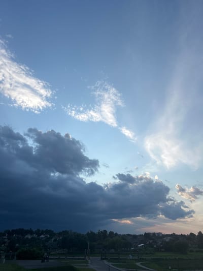 The image depicts a sky over a rural landscape, likely in the vicinity of Жухавіцкі сельскі Савет, Belarus. A dramatic contrast exists between the dark, ominous storm clouds gathering on the left and the lighter, scattered clouds and clear blue sky on the right, suggesting an approaching or departing weather front. Below the sky, a settlement with houses and trees is visible. The foreground shows a path or road with some fencing and greenery, indicating a developed area within the rural setting. There are no discernible people or specific actions taking place in the scene, which is a static, environmental depiction. The lighting suggests it is either early morning or late afternoon, given the interplay of light and shadow across the sky and land.