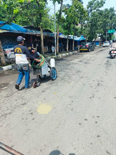 The image shows a man pushing a cart with a large spool of hose and a welding torch on it.  He is walking down a street in Cimahi, Indonesia, located at coordinates -6.893, 107.558. The street is lined with small shops and trees. There are several cars and motorcycles parked on the side of the road. The man is wearing a white shirt and blue jeans. The street is paved and looks to be in fair condition. There are several small, dark spots on the pavement, possibly oil. There is some debris on the street. The sky is clear and there is a blue car parked on the right side of the street. The man is looking forward and appears to be focused on his task.  There are no other people in the immediate vicinity.