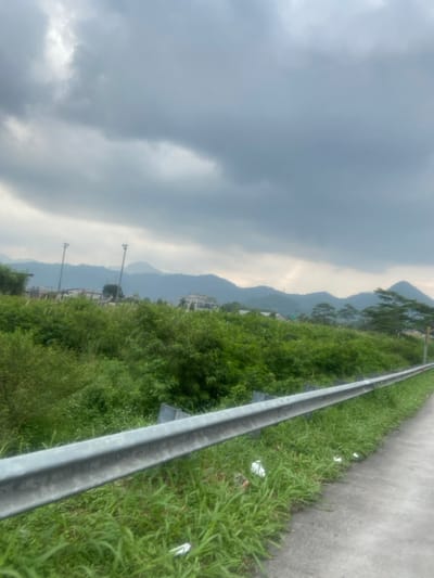 The image shows a view from a roadside in Margaasih, Indonesia, with a metal guardrail along a paved road. There is a dense growth of green foliage, shrubs and trees, in the foreground, and a row of hills in the distance. The sky is overcast with gray clouds. There are two tall, thin poles, possibly light posts, visible in the background. The image appears to have been taken on a cloudy, overcast day. The road is empty of traffic. The scene is typical of rural Indonesia, with the dense greenery and the rolling hills in the background.
