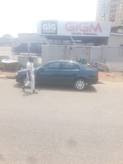 A man in a striped shirt and white pants walks on the right side of a dark green sedan. The car is parked parallel to a sidewalk in front of a building with a large sign that reads "GIG LOGISTICS" and "GIGM ENUGU TERMINAL". The sky is bright and overcast. The scene appears to be in Enugu, Nigeria.