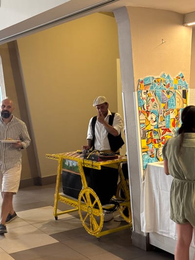 The image captures a scene inside a building, possibly a venue or a covered walkway, with two men and part of a woman visible.
On the left, a bald man with a beard is walking, holding a white plate with food. He is wearing a striped, short-sleeved shirt and light-colored shorts.
In the center, a man wearing a white shirt, a dark vest, and a light-colored cap is standing and appears to be interacting with a music setup on a yellow cart. The cart is decorated with colorful patterns and has a speaker on its front. A microphone and other electronic equipment are visible on top of the cart. The man is touching his chin, seemingly in thought.
On the right, the back of a woman wearing a green outfit is visible. She is standing next to a white tablecloth-covered surface, facing away from the camera. Behind her, a vibrant, mosaic-like artwork adorns the wall. The artwork is composed of numerous small, colorful tiles forming abstract shapes and figures.
The environment suggests an indoor event or gathering, with tiled flooring and cream-colored walls. The lighting appears to be artificial. The overall atmosphere seems casual and possibly festive. The presence of the music cart and the decorated wall hints at an entertainment or artistic element to the scene, potentially at an event in Capaci, Italy.